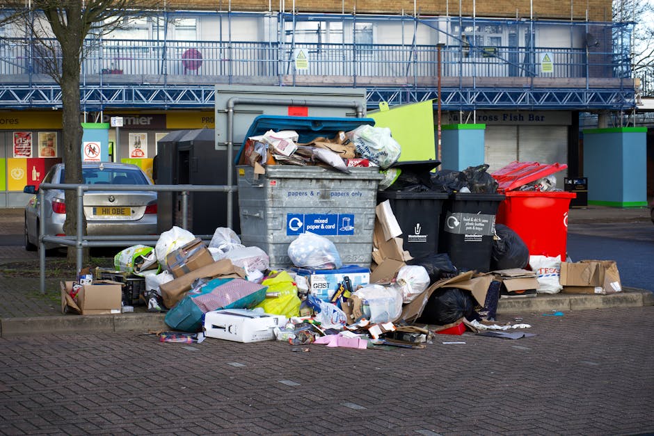 A large collection of overflowing rubbish bins and trash bags situated on a paved pavement in front of a commercial building, with various discarded packaging, cardboard boxes, plastic bags, and loose