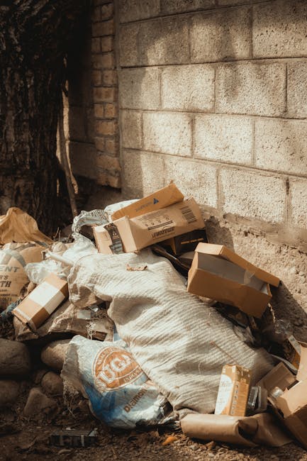 A pile of discarded cardboard boxes, some flattened and others partially intact, rests against a rough brick wall and a concrete block wall in an outdoor setting. The boxes vary in size, with some dis
